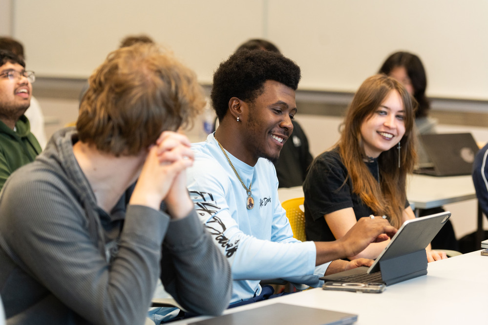 Students seated in a classroom use laptops and a tablet while participating in a group learning activity.