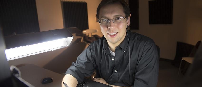 Person seated at a workstation uses a digital drawing tablet under bright task lighting in a studio or office environment.