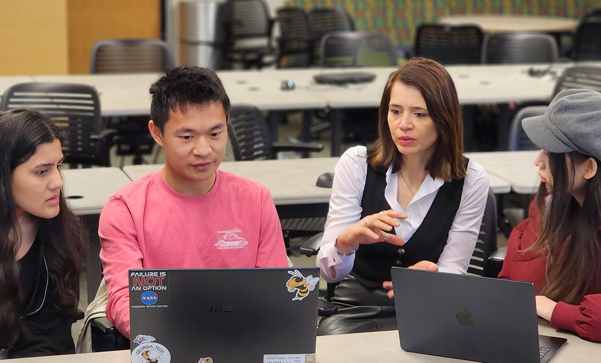 Four participants seated around laptops collaborate during a classroom or workshop session, discussing information displayed on their screens.