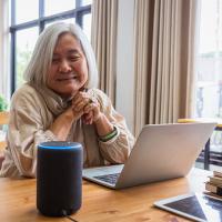 An elderly woman with short white hair smiles and looks at a smart speaker system.