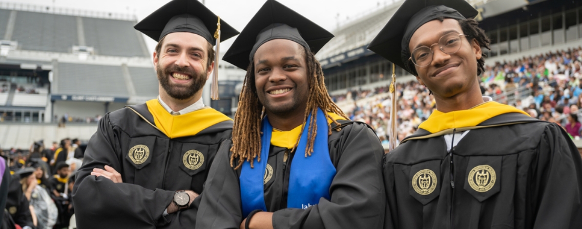 Three graduates wearing caps and gowns pose together in a stadium during a commencement ceremony.